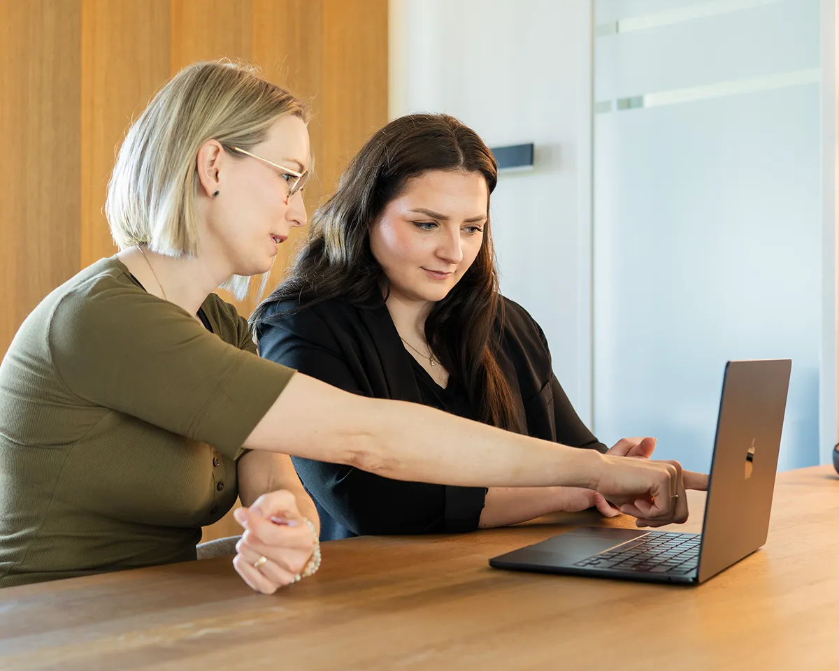 Zwei Frauen sitzen an einem Holztisch und schauen auf einen Laptop, wobei eine Frau auf den Bildschirm zeigt.
