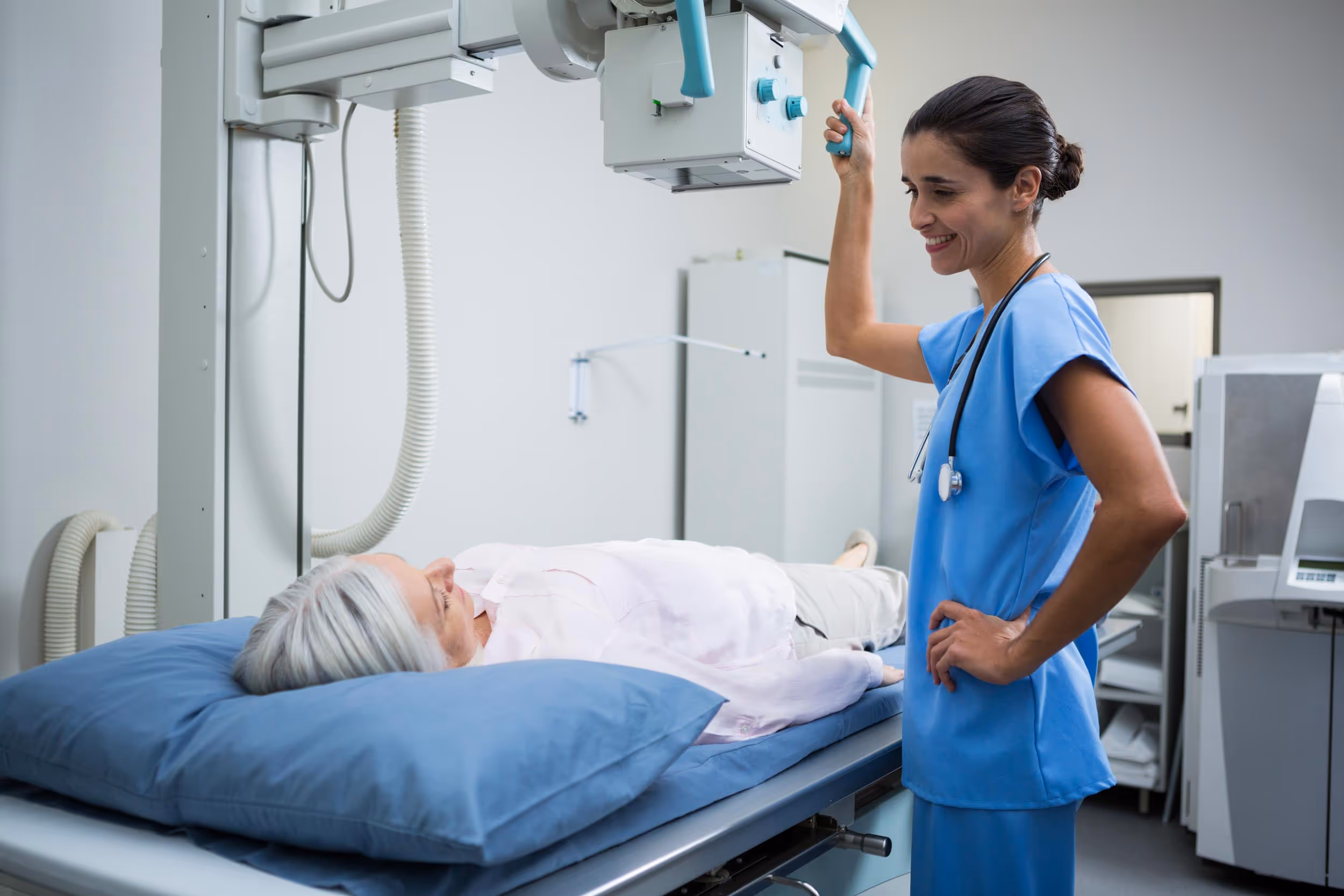 A nurse prepares an elderly woman for an X-ray while smiling and adjusting the machine.