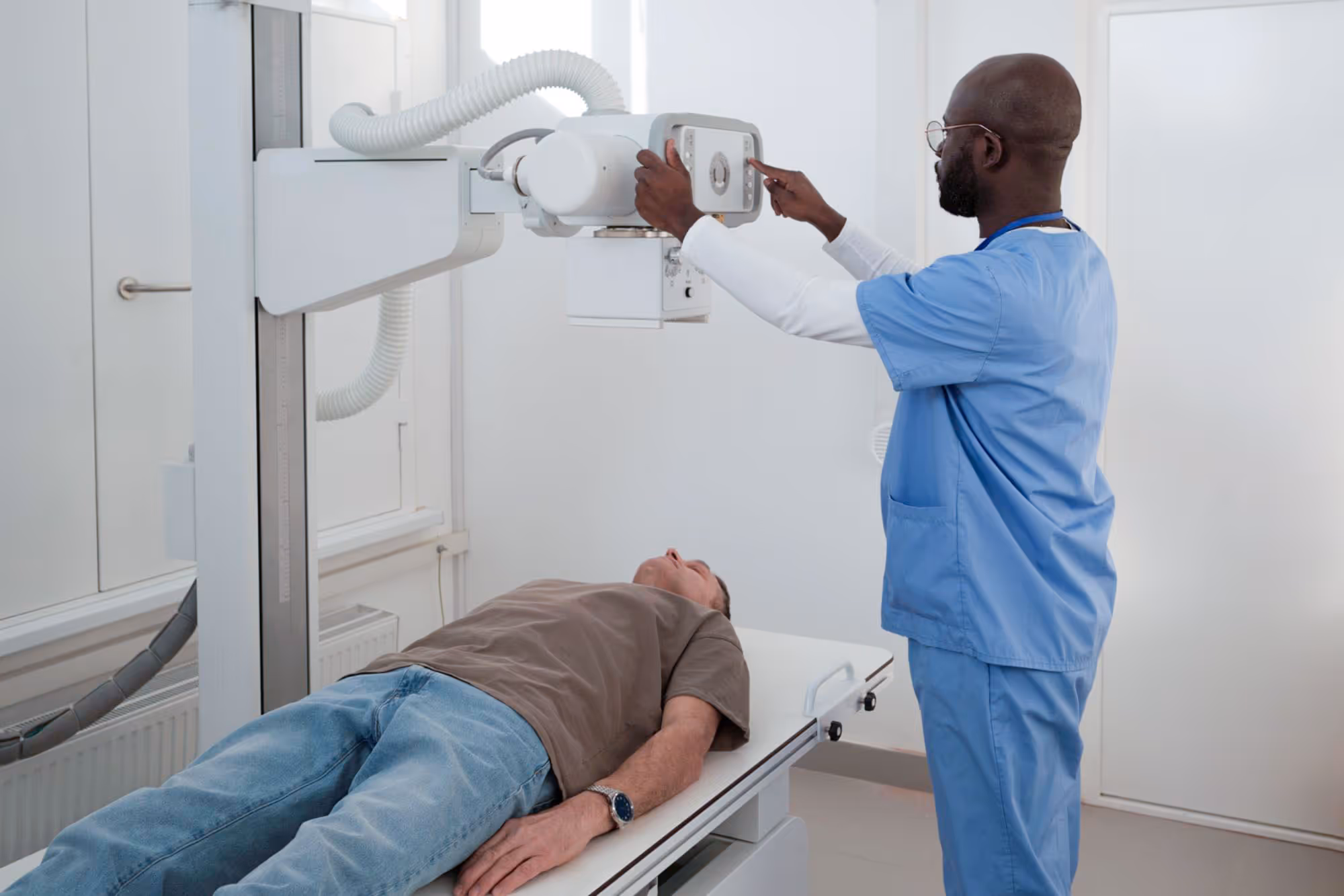 A healthcare worker in blue scrubs adjusts an X-ray machine above a patient lying down.