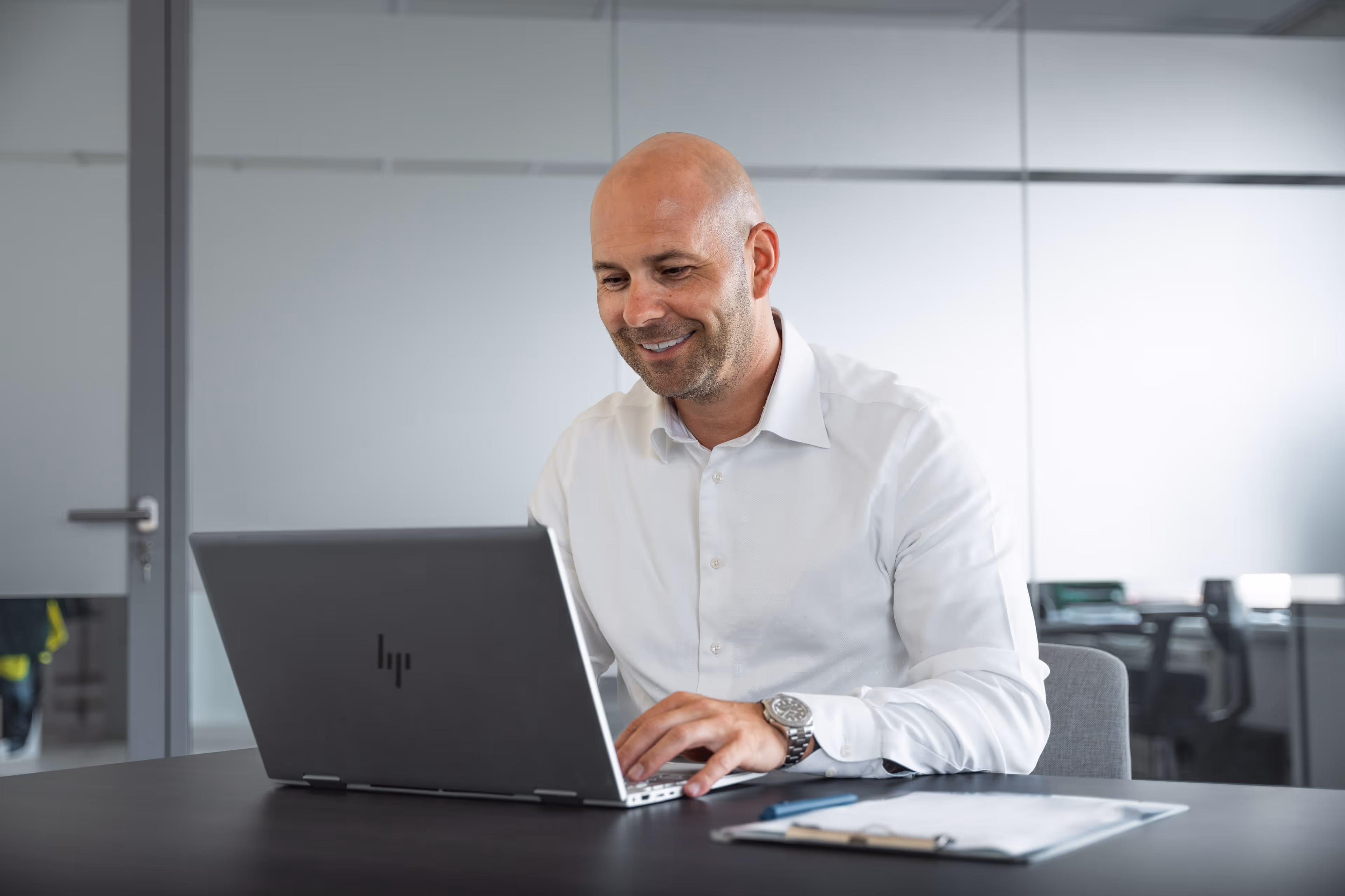 Man in witte hemd lacht terwijl hij werkt op zijn laptop aan een bureau.