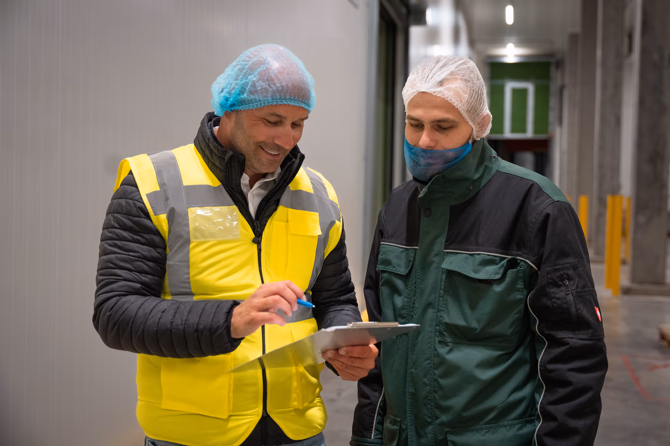 Twee mannen in beschermkledij bespreken documenten in een industriële omgeving.