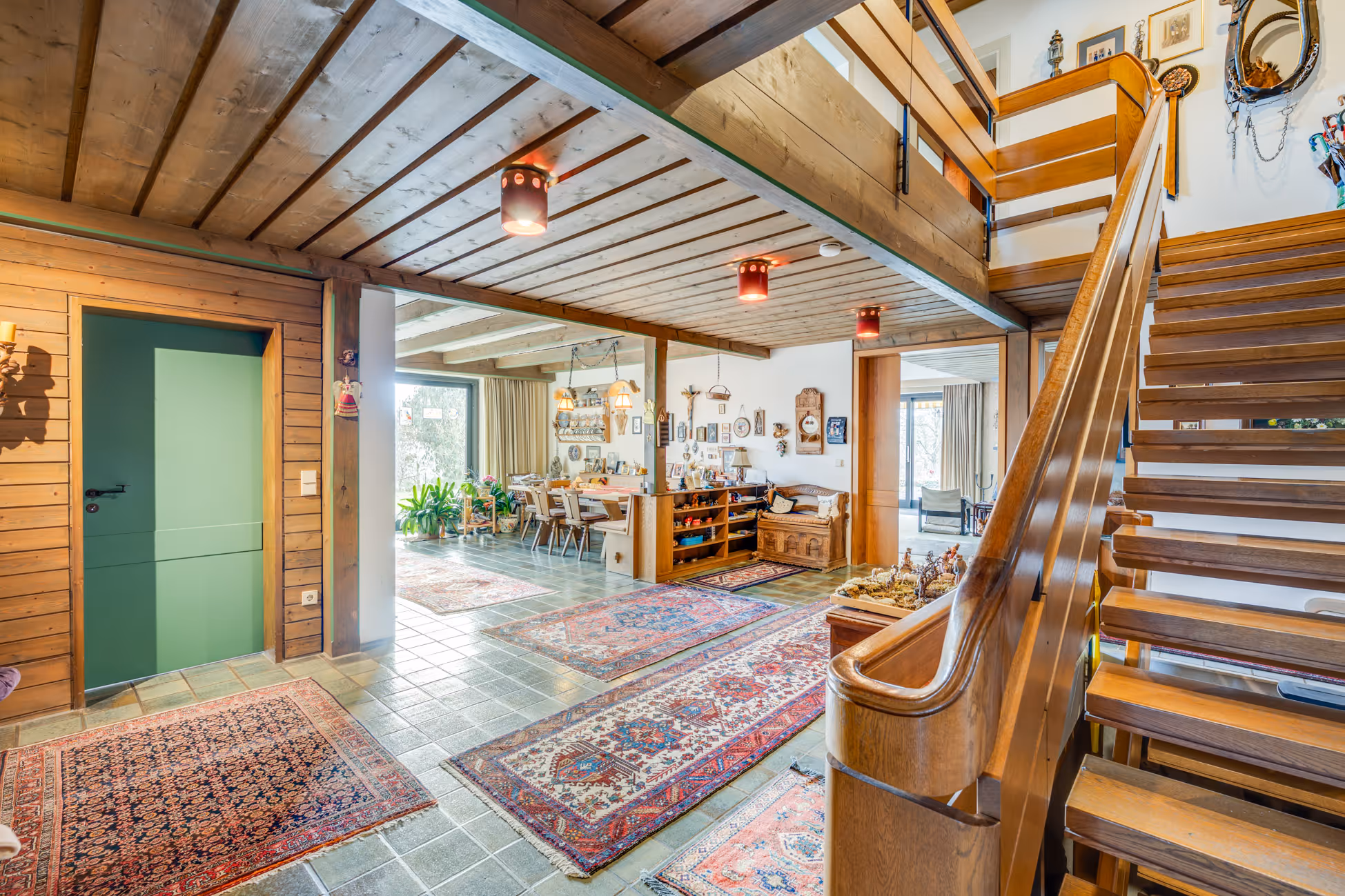 Wohnzimmer mit Holztreppe, Holzbalkendecke, mehreren persischen Teppichen und einem Essbereich mit Holzmöbeln.