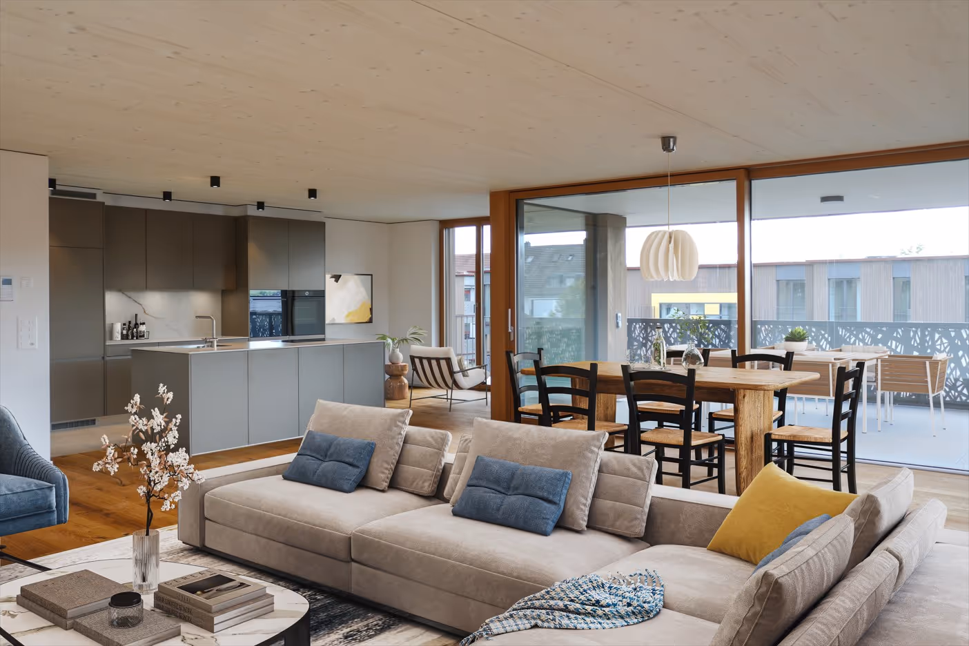 Modernes offenes Wohnzimmer mit beigem Sofa, blau-grauen und gelben Kissen, Essbereich mit Holztisch und schwarzen Stühlen, und integrierter Küche mit grauen Schränken.