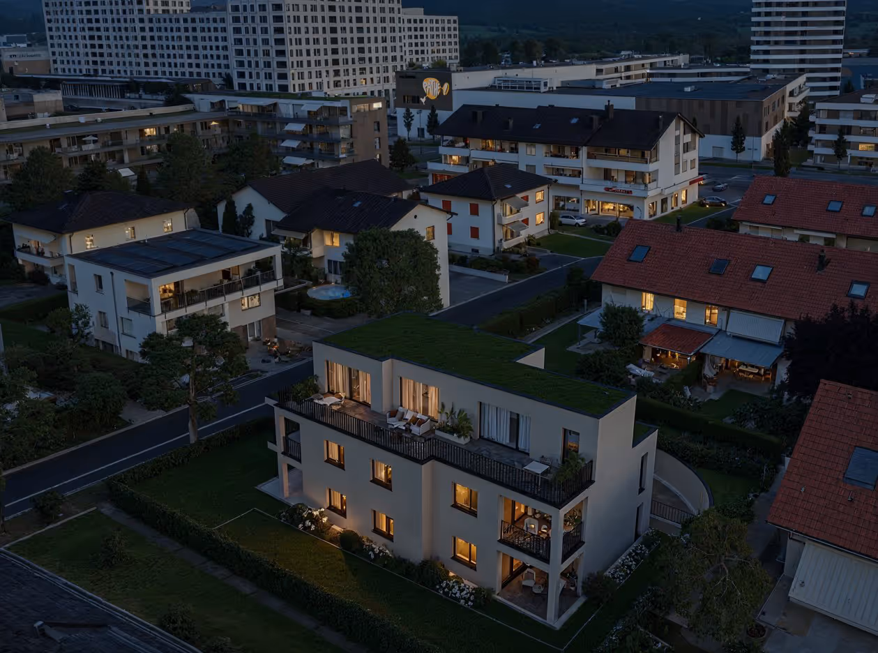 Abendaufnahme eines modernen Wohngebäudes mit begrüntem Dach und beleuchteten Fenstern in einem ruhigen Viertel.