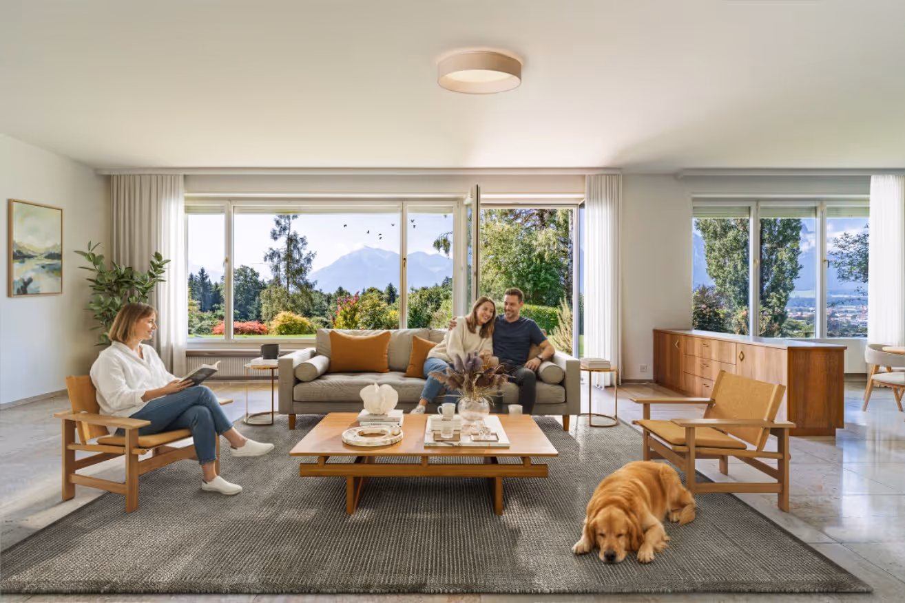 Heller Wohnzimmer mit großen Fenstern, Paar sitzt auf Sofa, Frau liest Buch auf Holzstuhl, Hund liegt auf grauem Teppich.