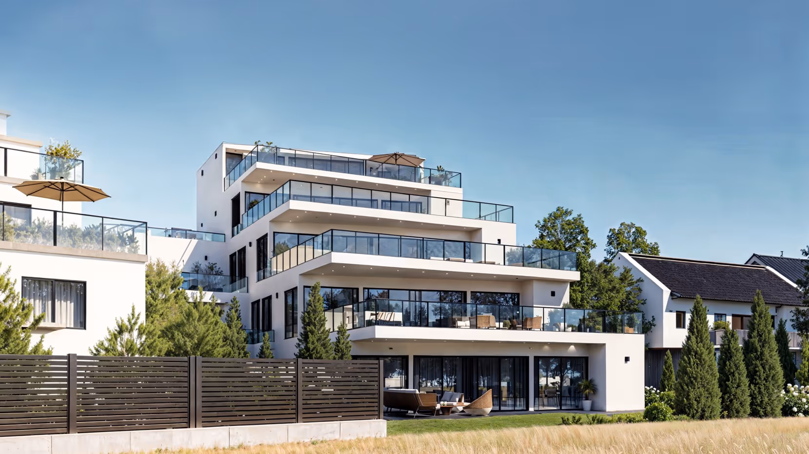 Modernes mehrstöckiges Wohnhaus mit großen Glasbalkonen und Gartenmöbeln, umgeben von grünen Bäumen und Hecken.
