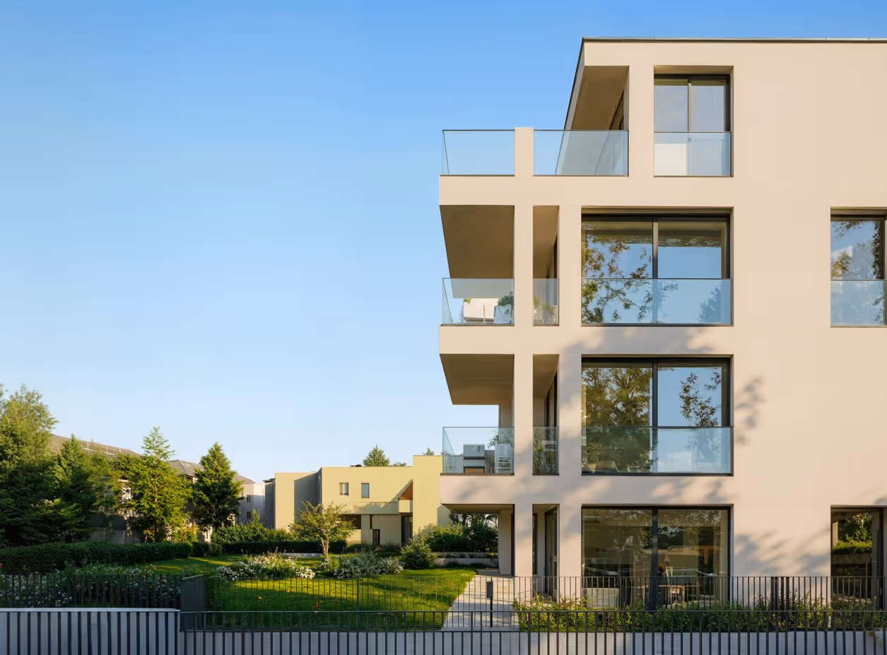 Modernes Wohngebäude mit großen Fenstern und Glasbalkonen, umgeben von grünen Pflanzen und Bäumen unter klarem blauem Himmel.