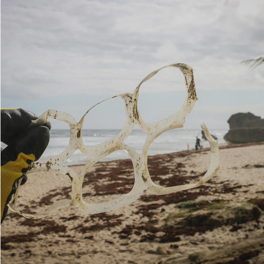 A hand holding up some plastic rings that are used to store cans. The image is set on the beach
