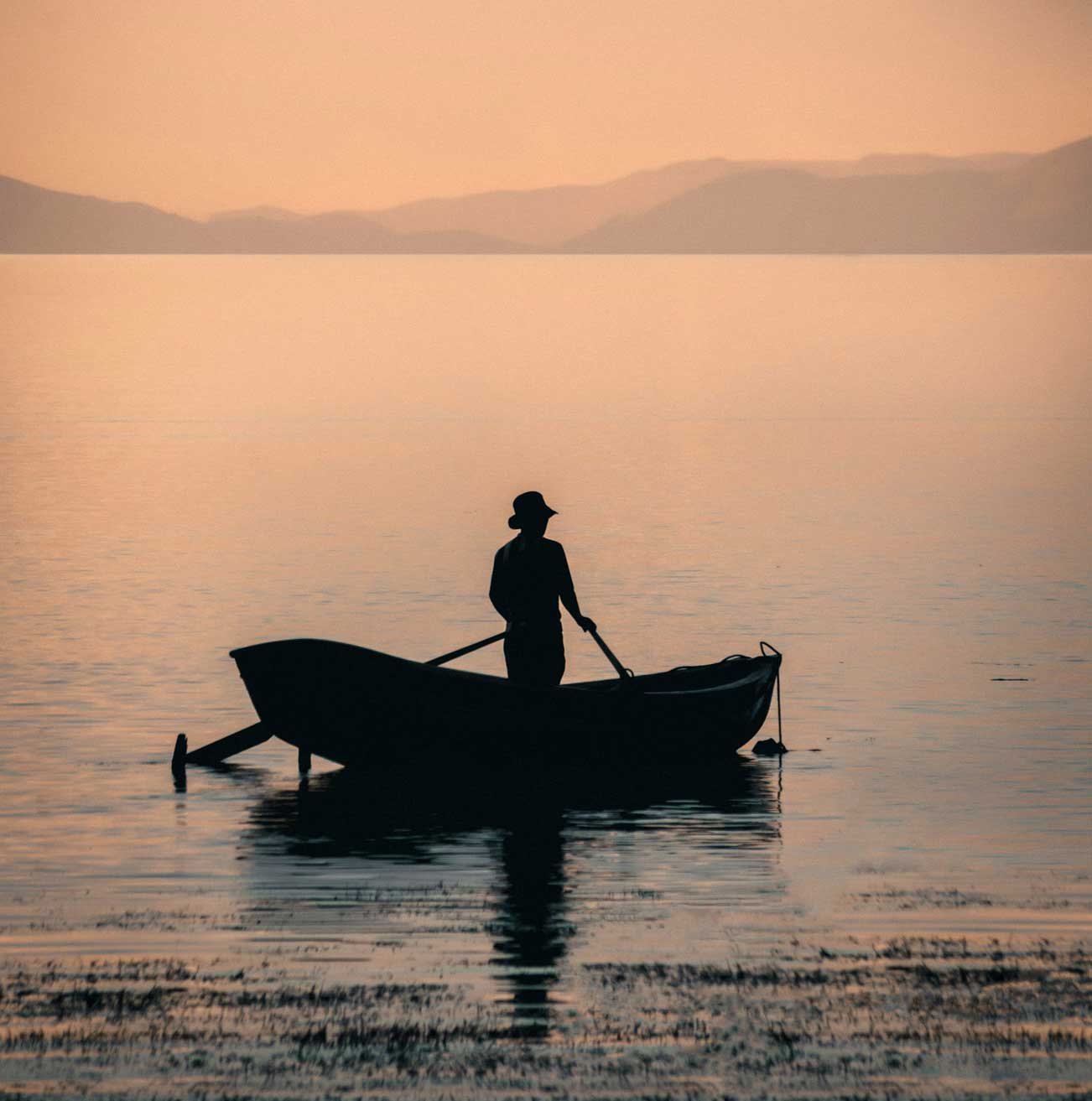 Silhouette of a person rowing a boat on calm water during sunset with distant mountains in the background.