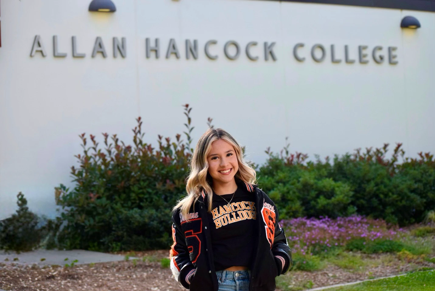 A student wearing a black varsity jacket stands in front of the Allan Hancock College sign, surrounded by green shrubs and flowers.