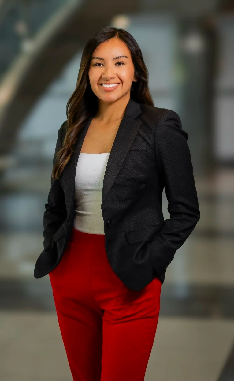 A professional woman in a black blazer and red pants poses confidently in a modern indoor setting.