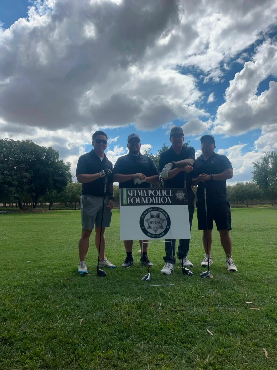 Four individuals in dark polo shirts stand on a golf course, holding clubs beside a sign for the Selma Police Foundation under a cloudy sky.