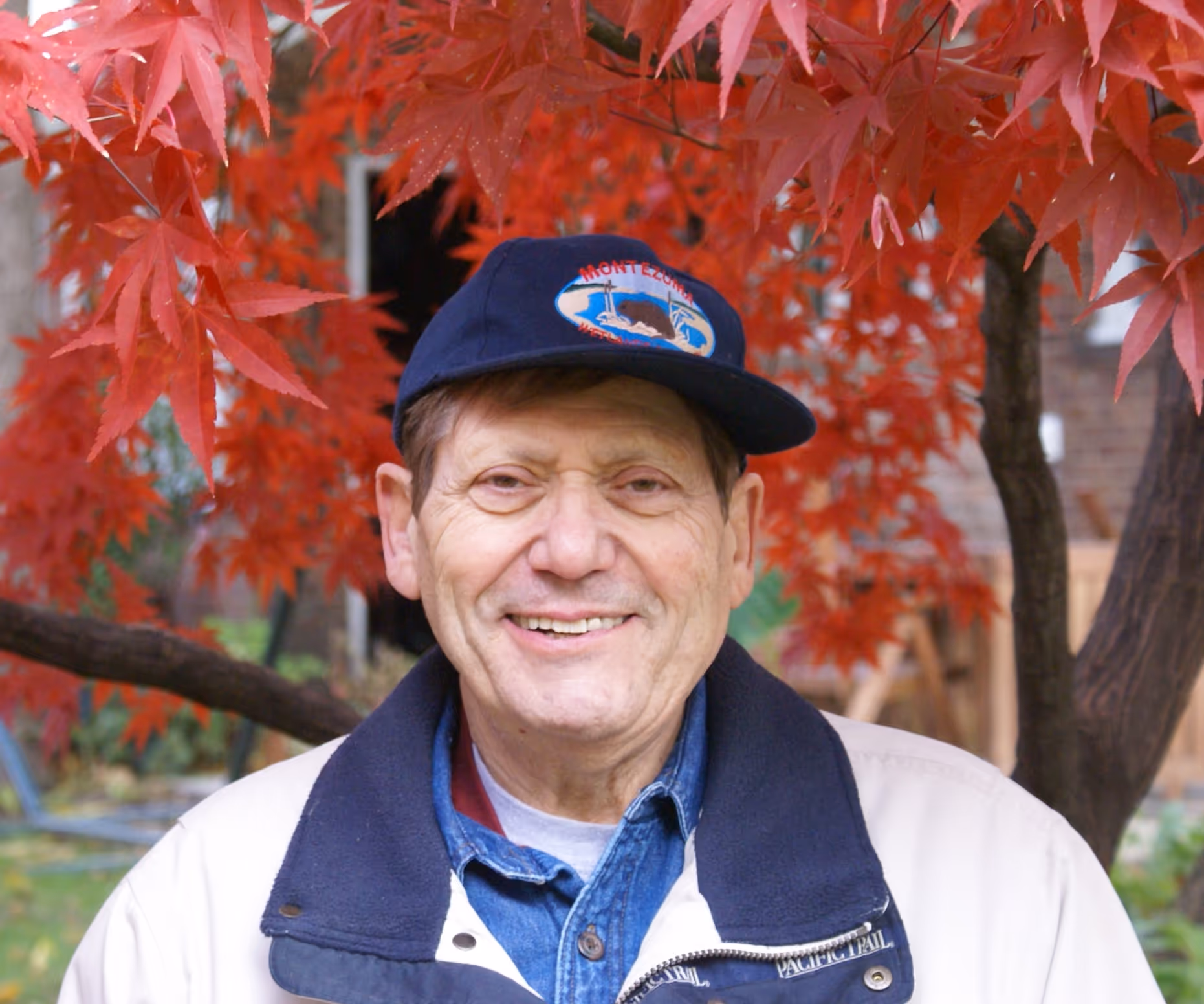 A male person in a blue hat, smiling with a tree with red leaves in the background.