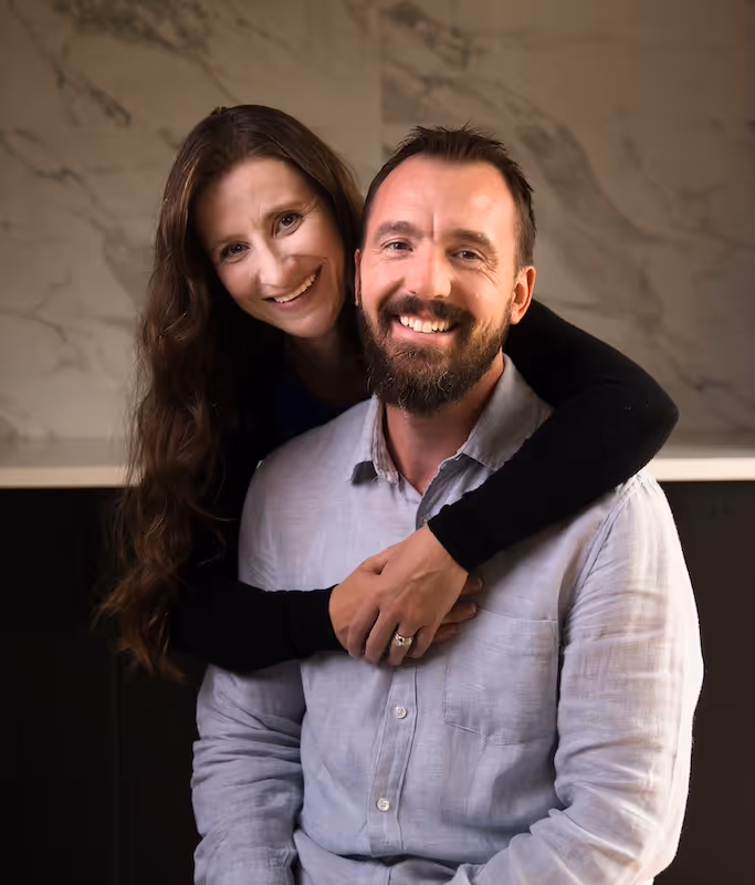 Smiling woman with long brown hair hugging a bearded man in a light gray shirt from behind against a marble wall background.