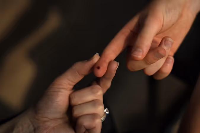 Close-up of two hands touching fingertips, one fingertip with a small drop of blood.