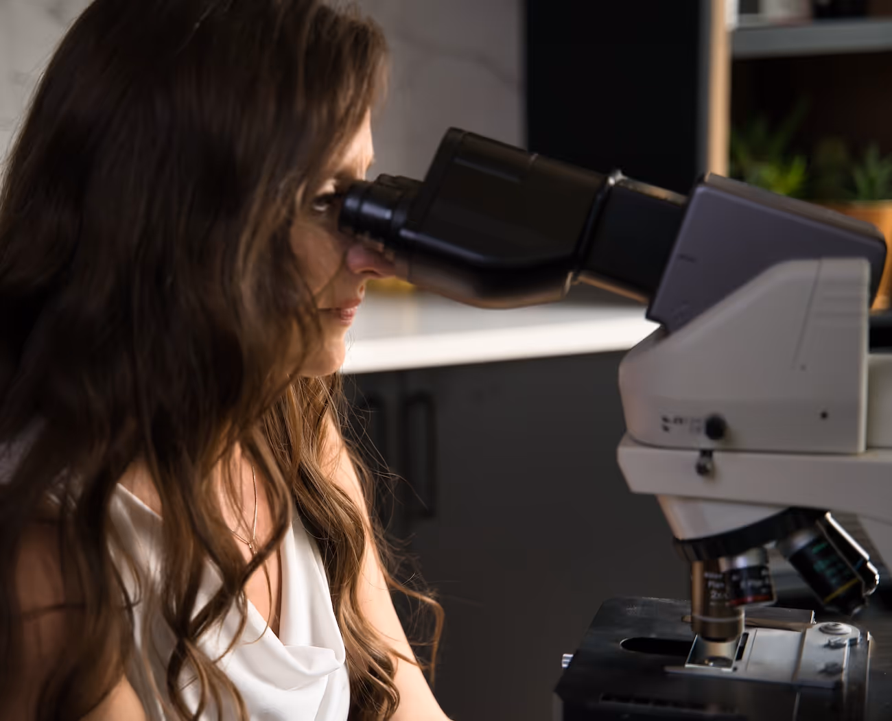 Woman with long hair looking through a microscope in a lab setting.