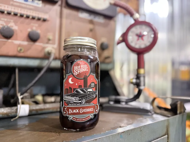 Glass jar labeled Hot Rod Shine Black Cherries filled with dark cherries flavored moonshine on a metal surface.