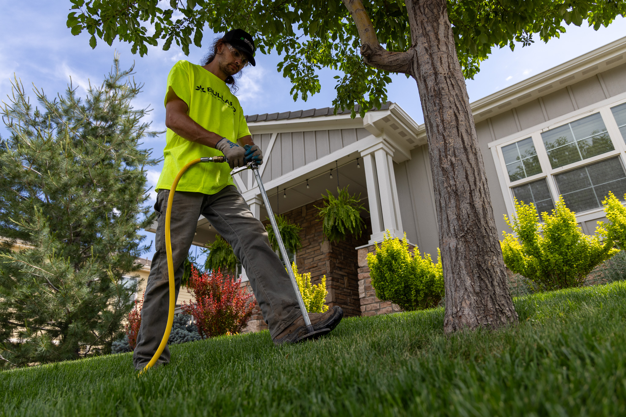 Tree service professional using specialized equipment for tree care in a well-maintained residential garden. The worker wears a bright yellow shirt with Bullard Tree & Garden branding, indicating expertise in Boulder's tree service industry. The scene includes a healthy lawn, a mature tree, and a house with attractive landscaping.
