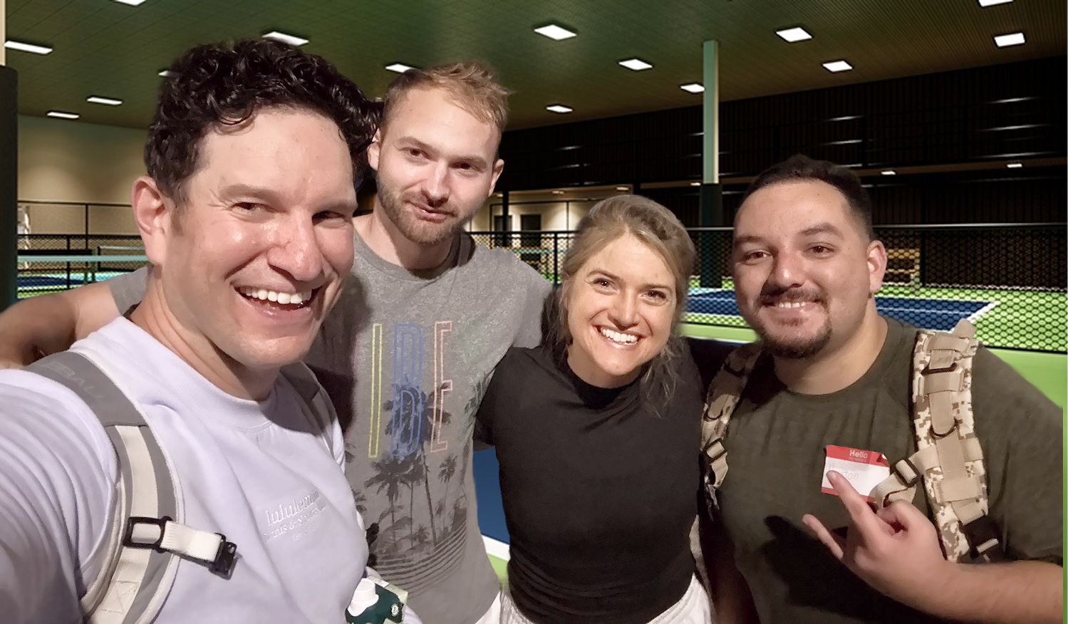 Group of four young adults smiling and posing for a selfie indoors near a fenced sports court.