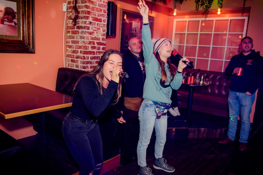 People singing karaoke in a dimly lit room with microphones, one person with arm raised, others watching and enjoying.