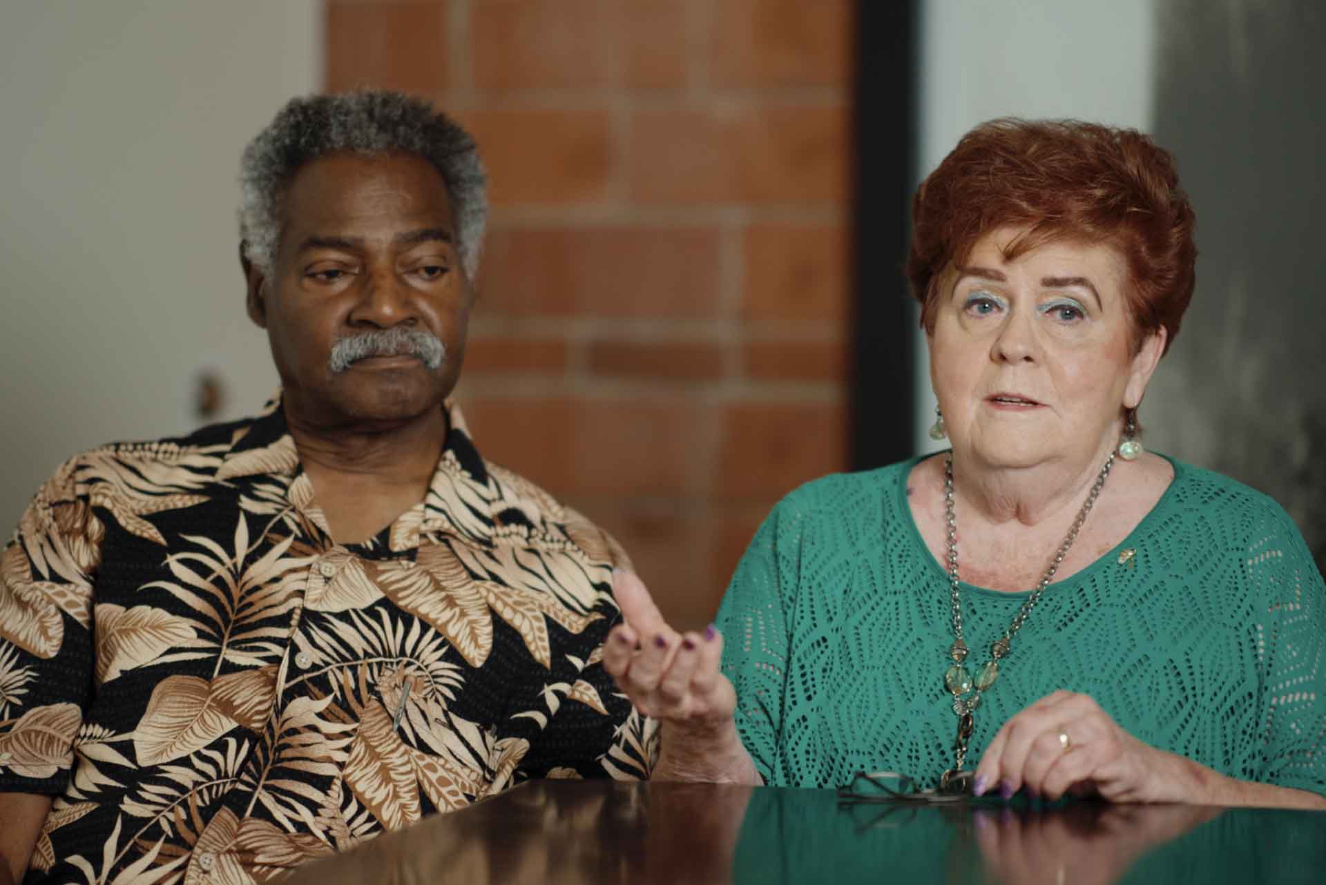 Older man in patterned shirt and woman in teal top talking while seated at a table indoors.