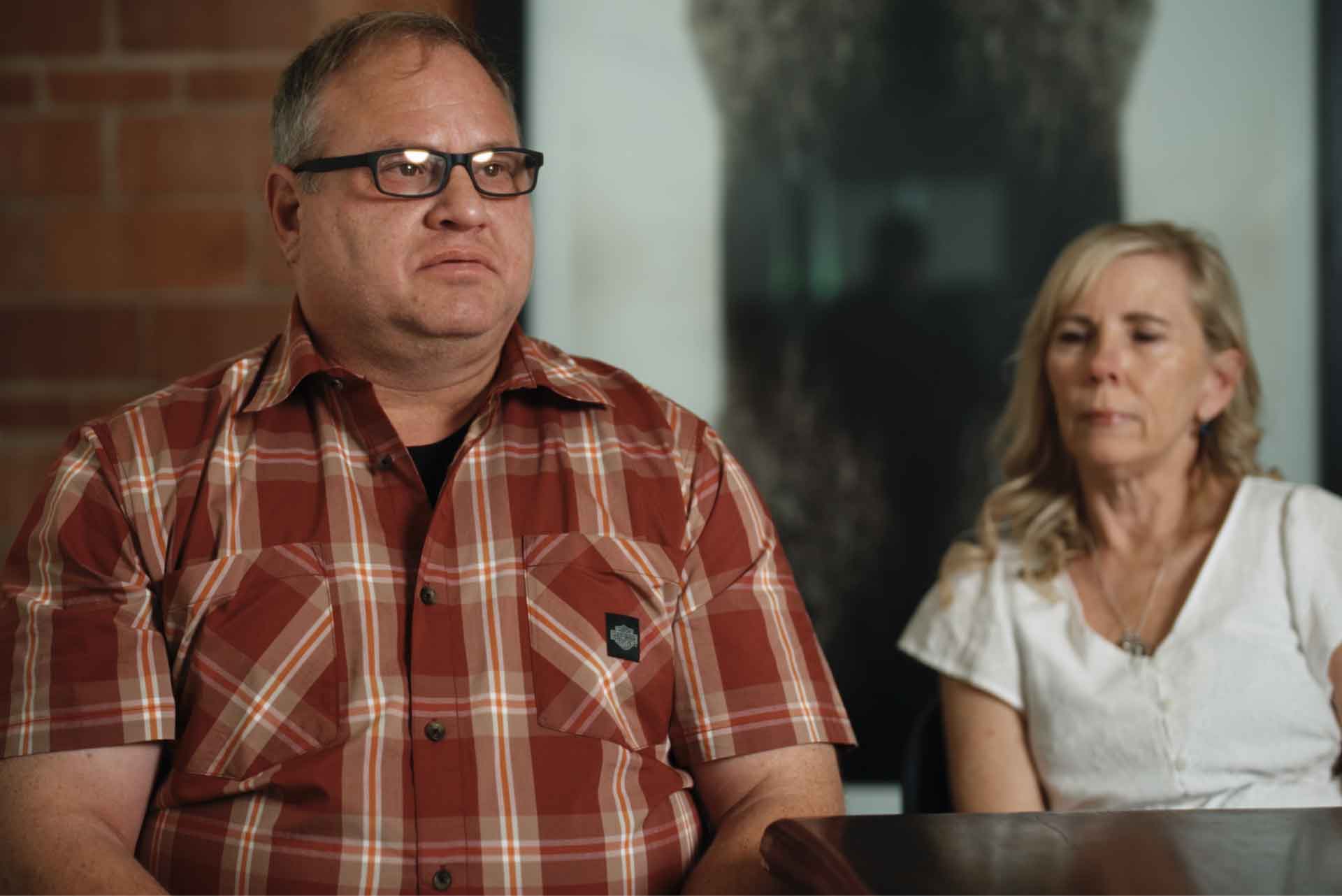 Man in glasses and red plaid shirt sitting next to a blonde woman in a white blouse at a table.