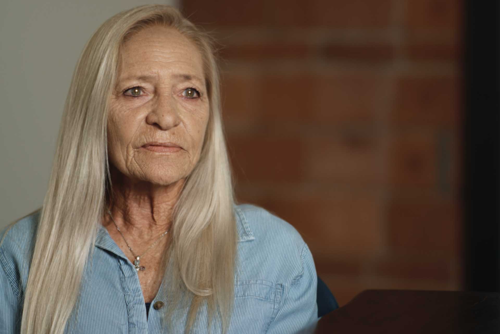 Older woman with long blonde hair wearing a light blue shirt and a cross necklace looks off camera.