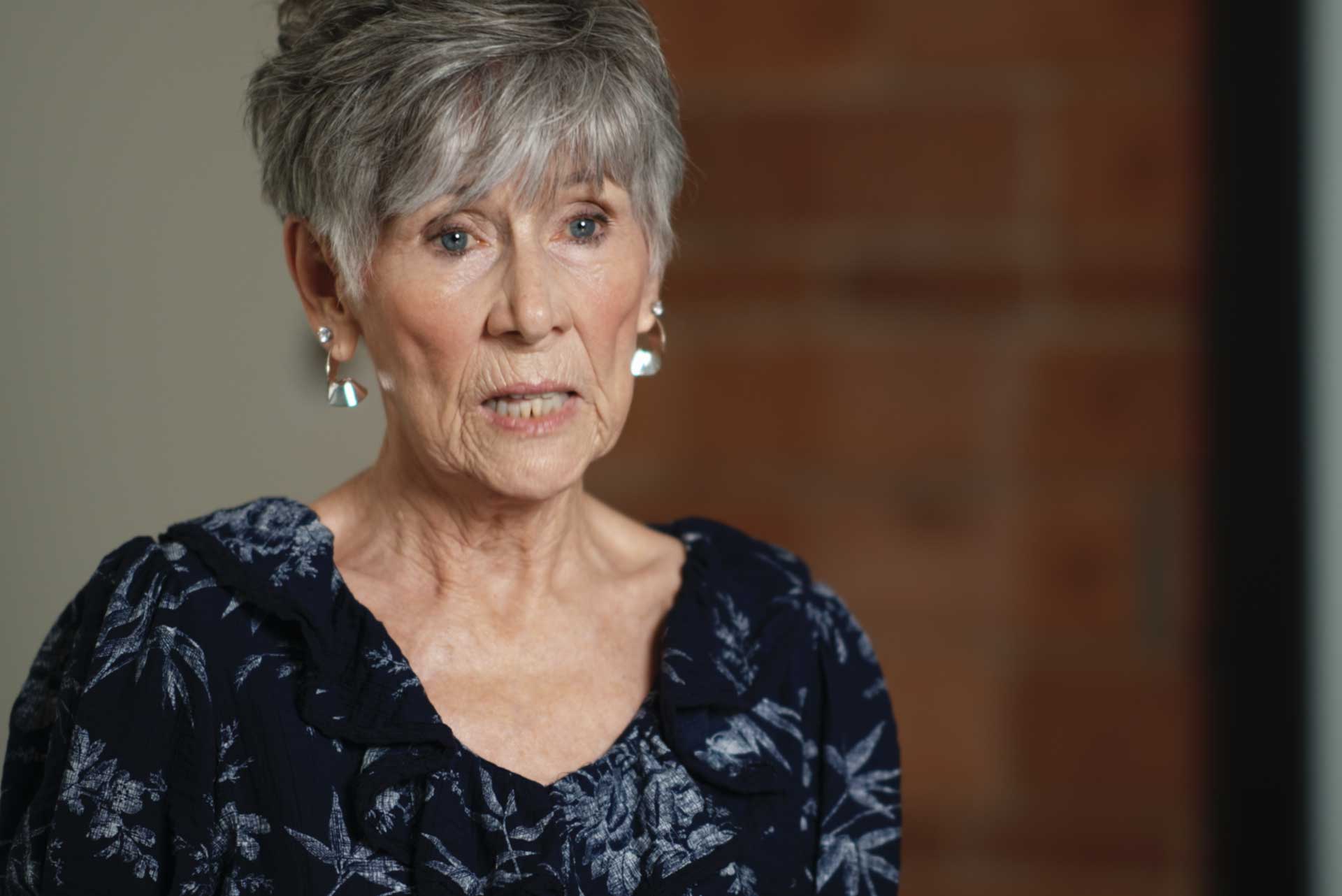 Older woman with short gray hair and earrings wearing a dark floral blouse against a blurred background.