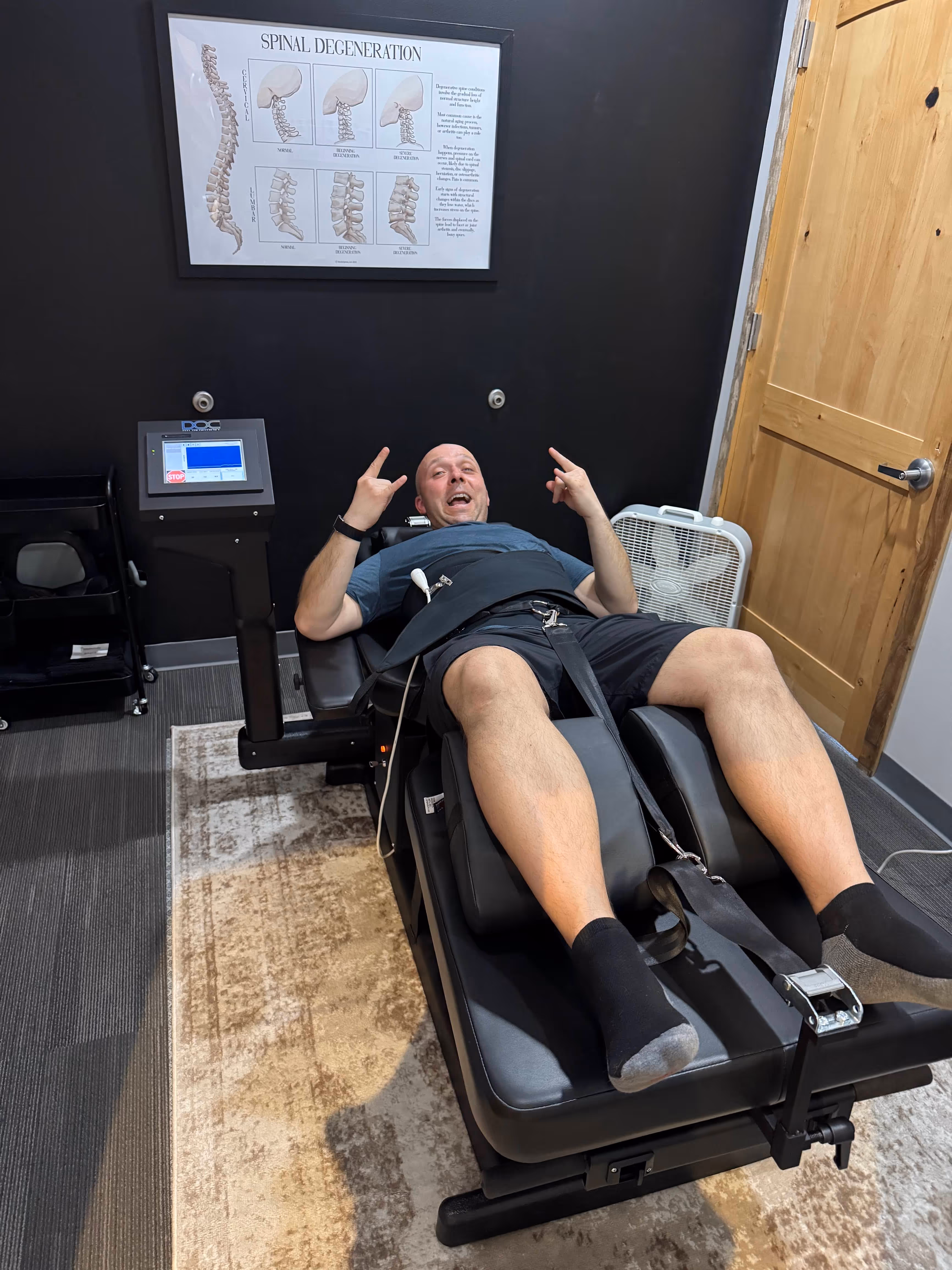 Patient smiling and giving rock-on hand gestures while lying on a spinal decompression table at Elite Wellness Chiropractic, with a spinal degeneration chart on the wall behind him