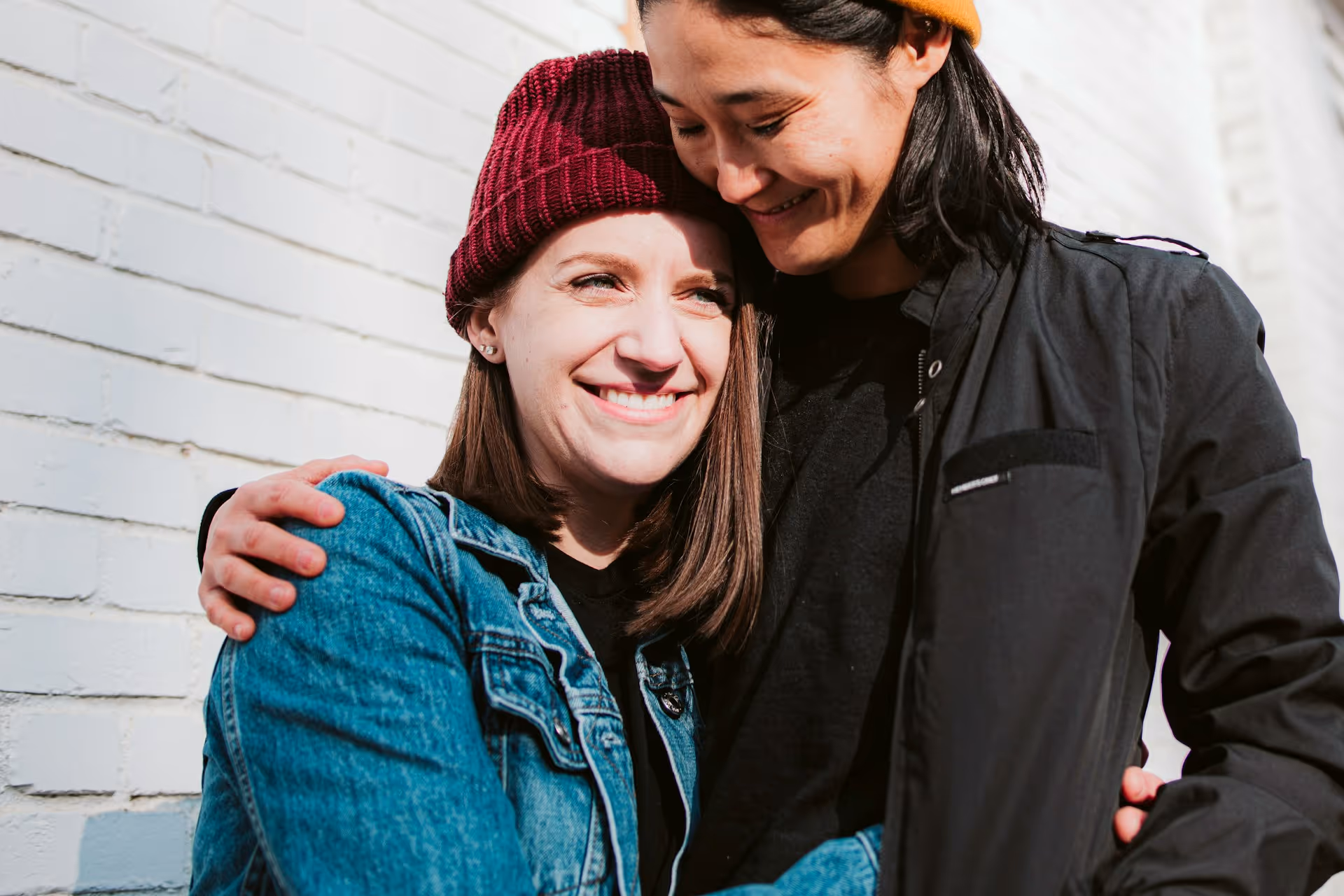 Home is about who you share it with. Two people in casual winter clothing embracing and smiling together in front of a white brick wall.