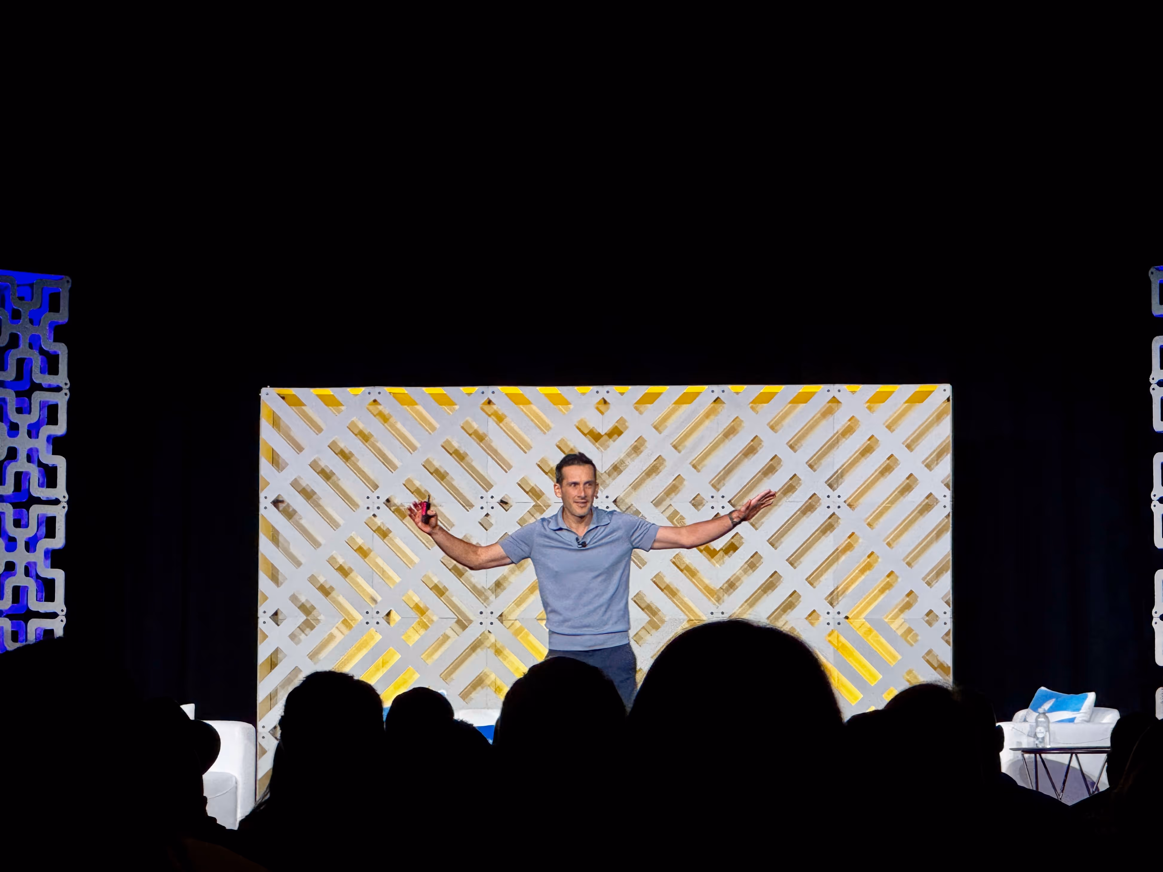 Dave Meyer stands on stage with arms outstretched, facing an audience at his BPCON keynote. He is in front of a yellow-and-white geometric background; silhouettes of real estate investors can be seen in the foreground.