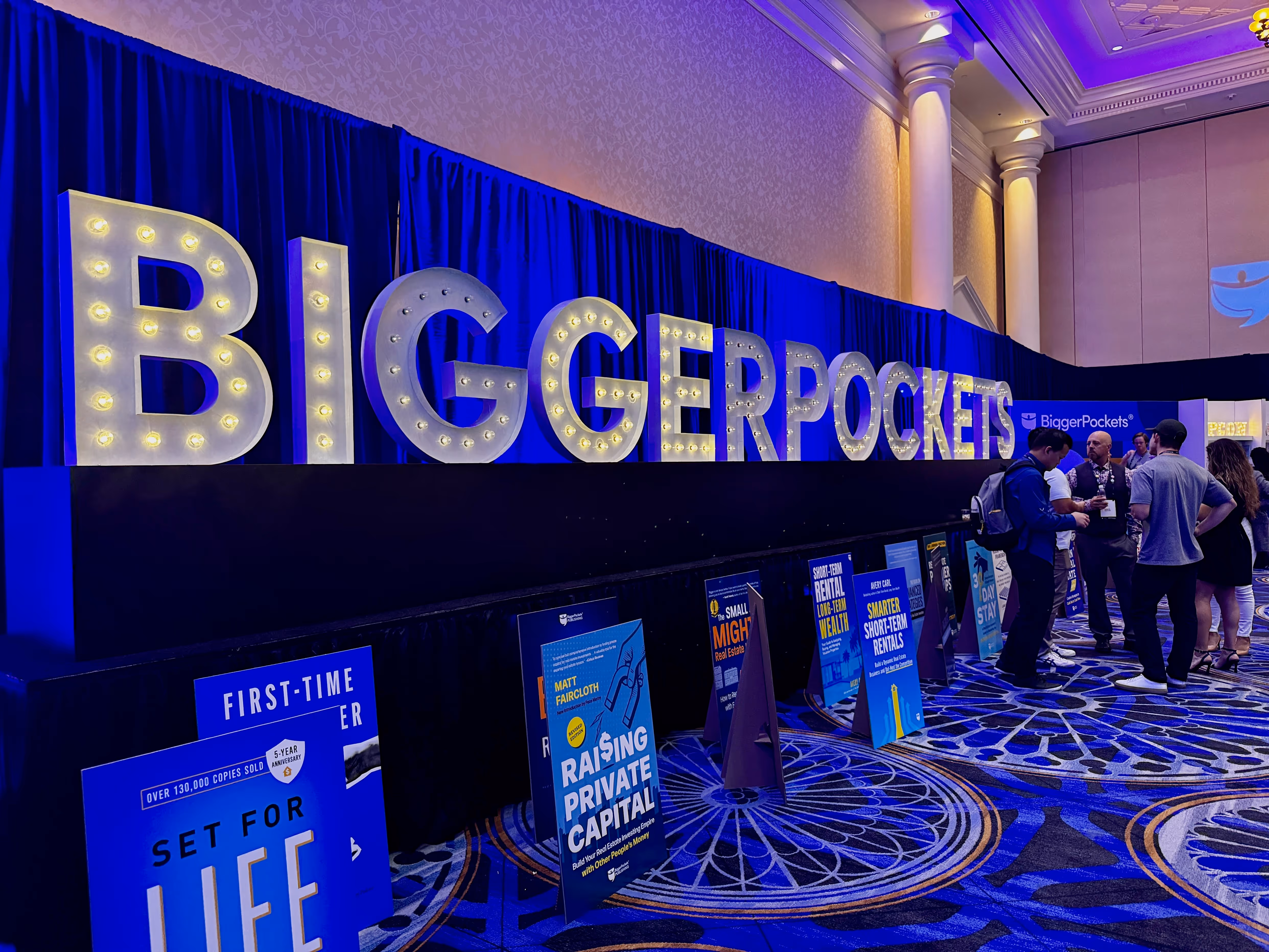 A large, illuminated "BiggerPockets" sign shines in the BPCON exhibit hall, where real estate investors gather near book displays and posters. Elegant columns and patterned carpet set the scene at BPCON 20265