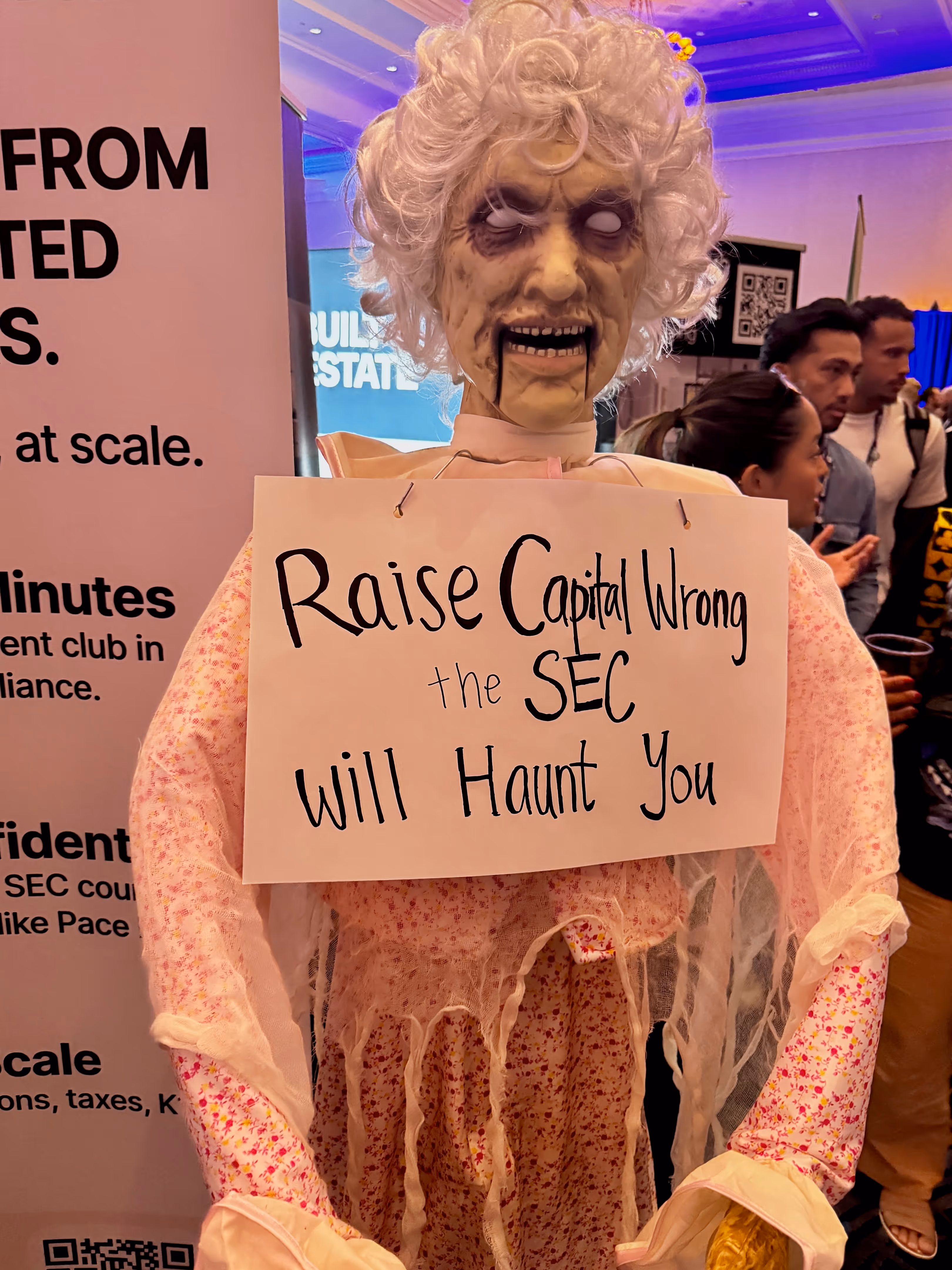A zombie mannequin in a pink nightgown and white wig holds a sign reading, "Raise Capital Wrong the SEC Will Haunt You," at BPCON 2025.