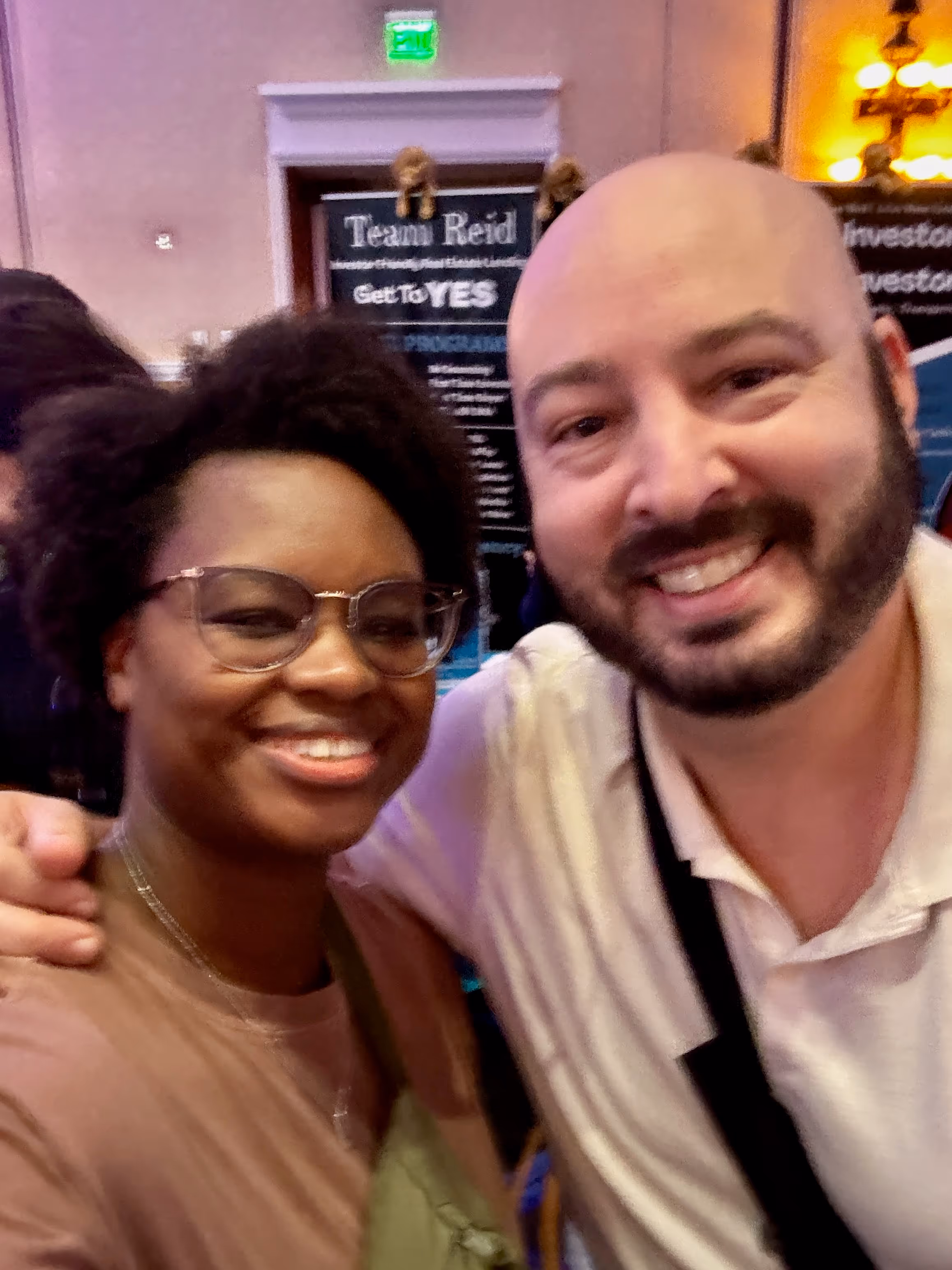 Brittany, Joint Founder of Our Neighborhood, with David Greene, smiling and posing for a selfie at BPCON 2025. The background shows informational posters for real estate investors and soft lighting. Both individuals appear happy and friendly, enjoying the BPCON sessions.