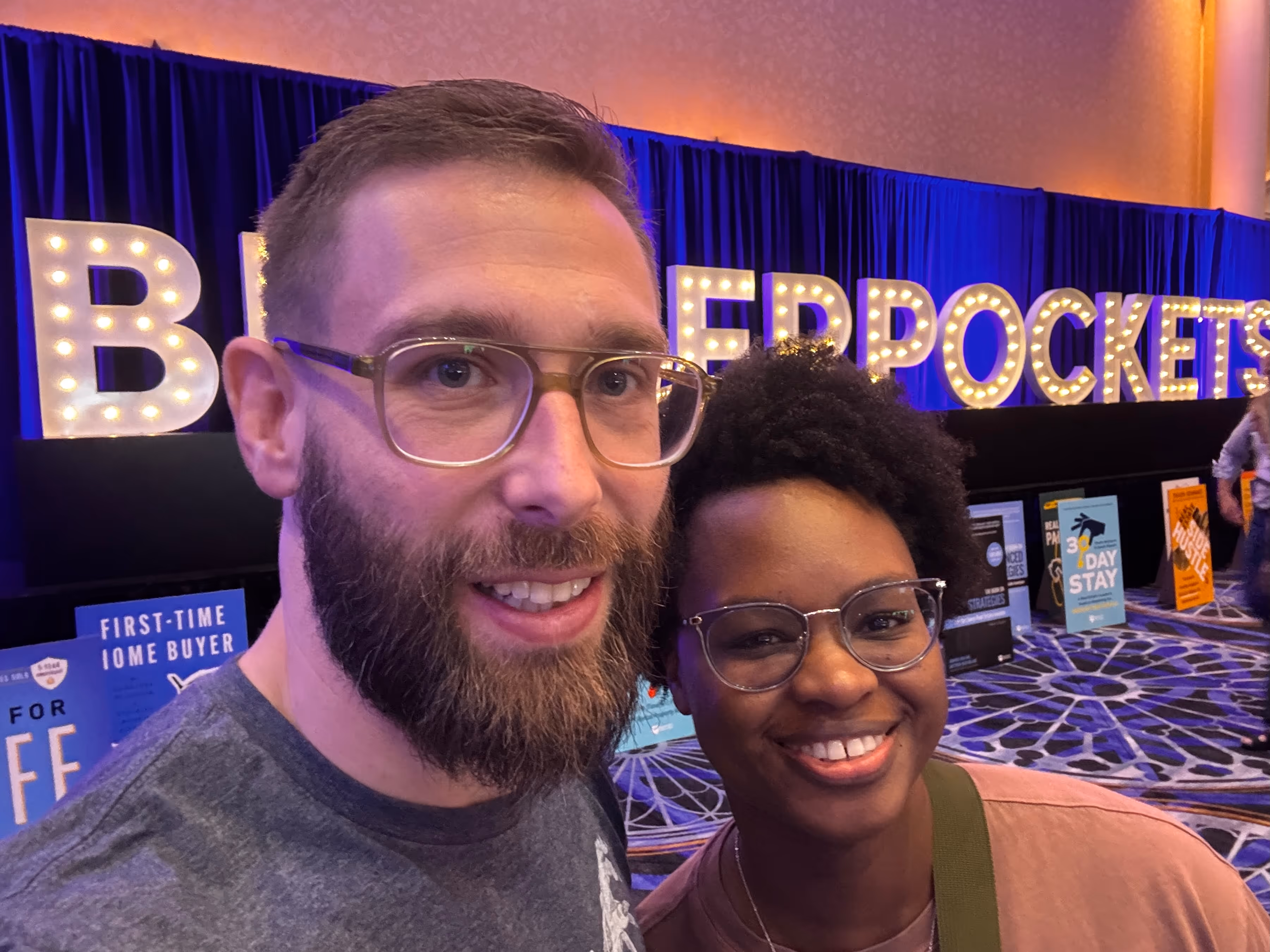 Brittany and Sebastian, Joint Founders of Our Neigborhood, smiling in front of illuminated "BIGGERPOCKETS" letters at BPCON 2025, an event attracting real estate investors and featuring informational posters.