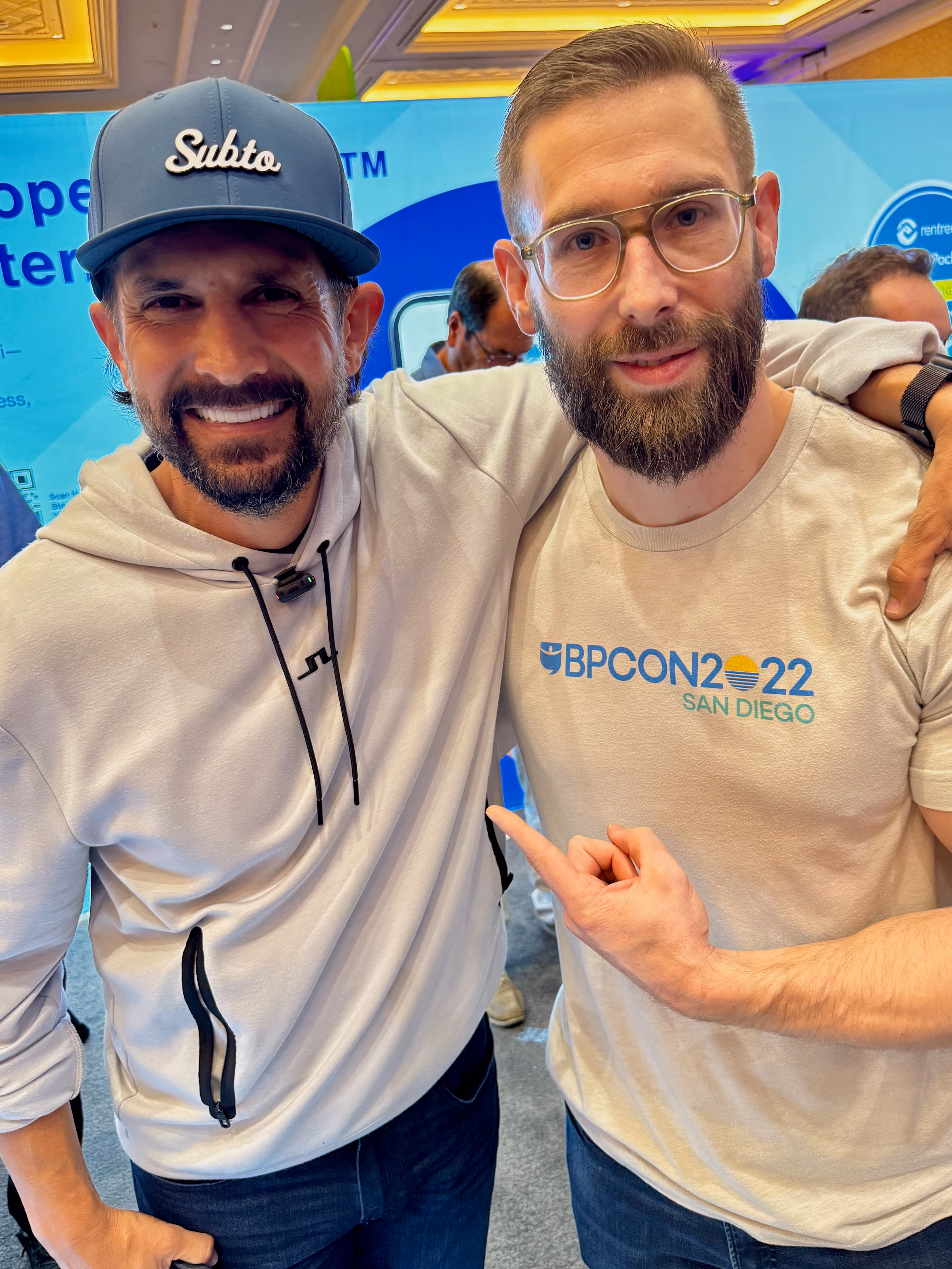 Sebastian, Joint Founder of Our Neighborhood, and Pace Morby pose together in front of the RentRedi exhibit hall booth at BPCON 2025. One wears a white hoodie and navy cap, the other sports glasses and a BPCon 2022 San Diego t-shirt while pointing at it.