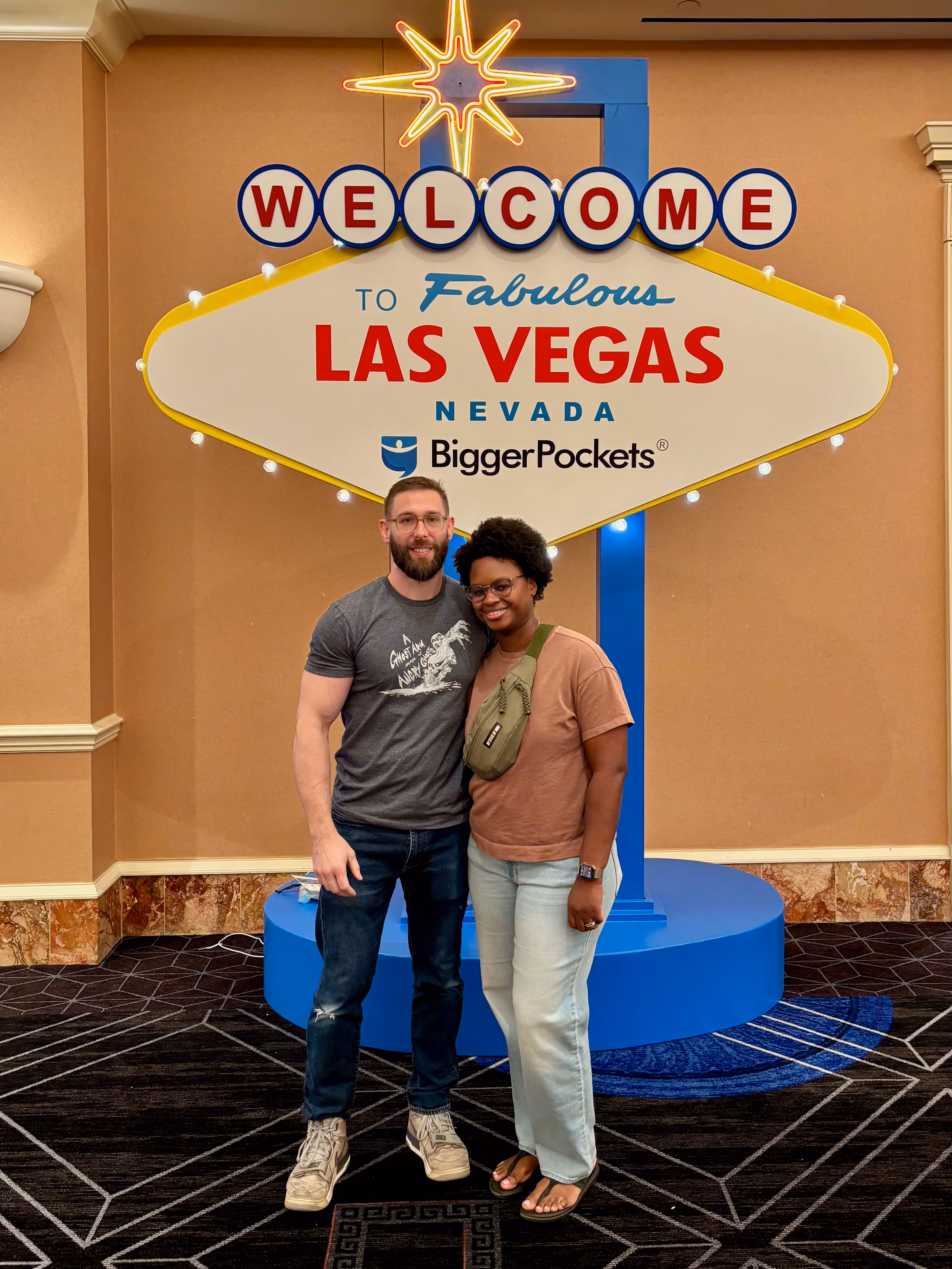 Brittany and Sebastian, Joint Founders of Our Neigborhood, smile and pose in front of a large "Welcome to Fabulous Las Vegas, Nevada" sign with "BiggerPockets" branding, celebrating BPCON 2025 inside a carpeted event space.