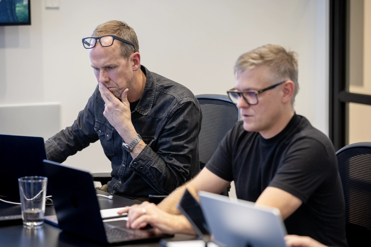 Two 303 Software developers focused on their laptops during a working session, one thinking through a problem while the other works through code.