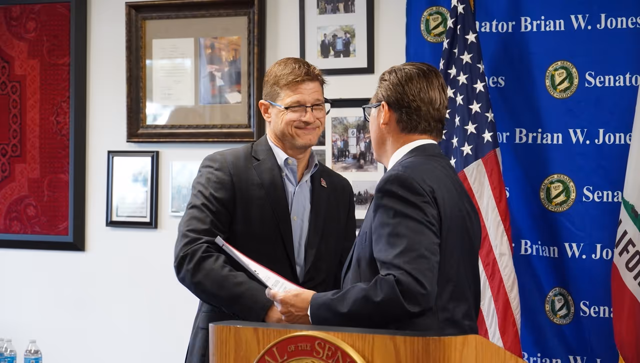 Two men shake hands in front of a flag.
