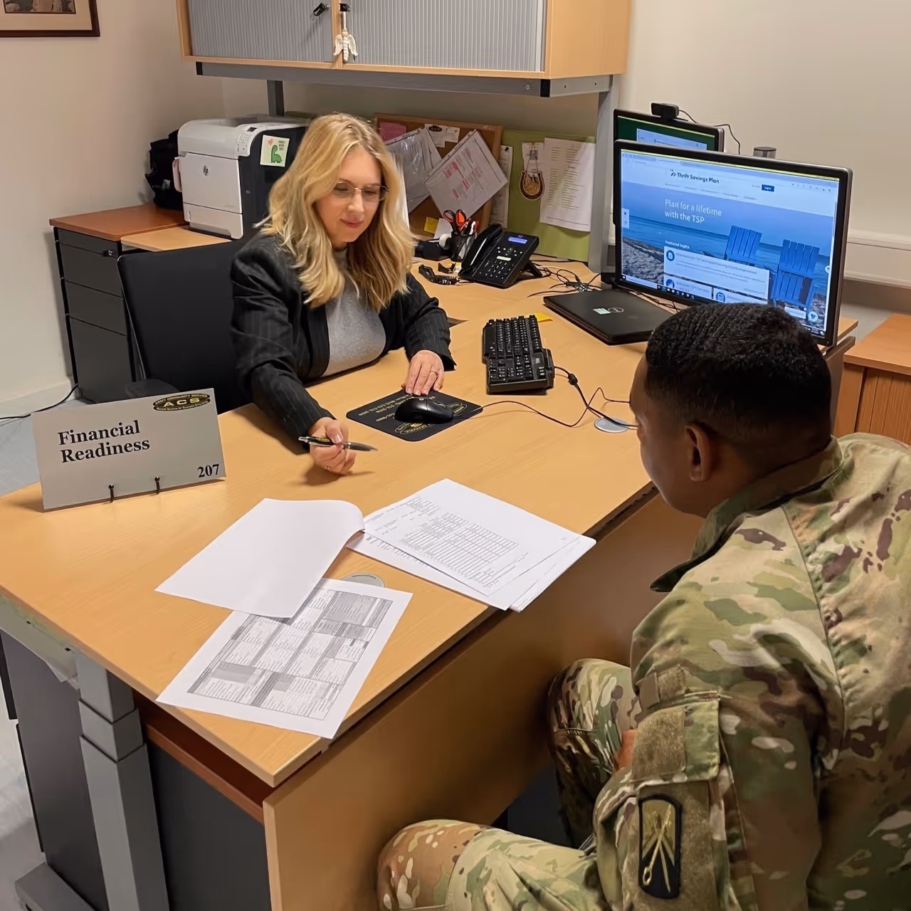 A woman and a man sit at a desk with papers and a computer.