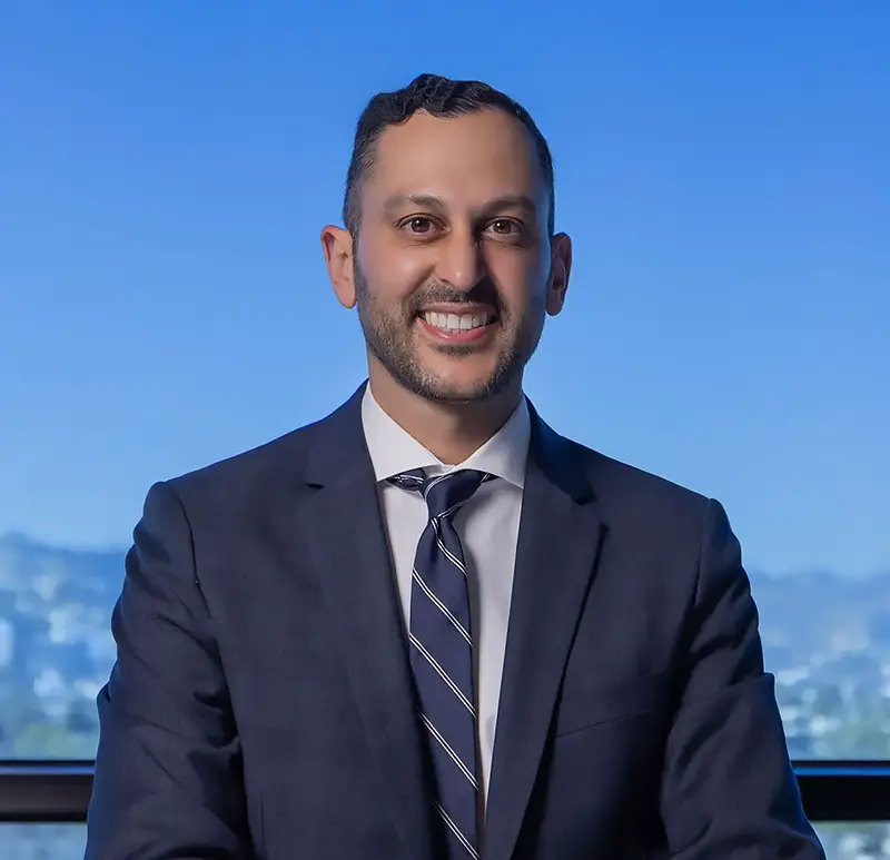 Dr. Yashar in a dark suit sitting in front of a cityscape and blue sky background.