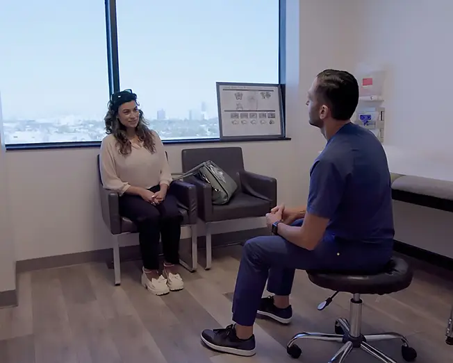 A woman sitting and talking with a male doctor in a medical office with large window and medical chart on the wall.