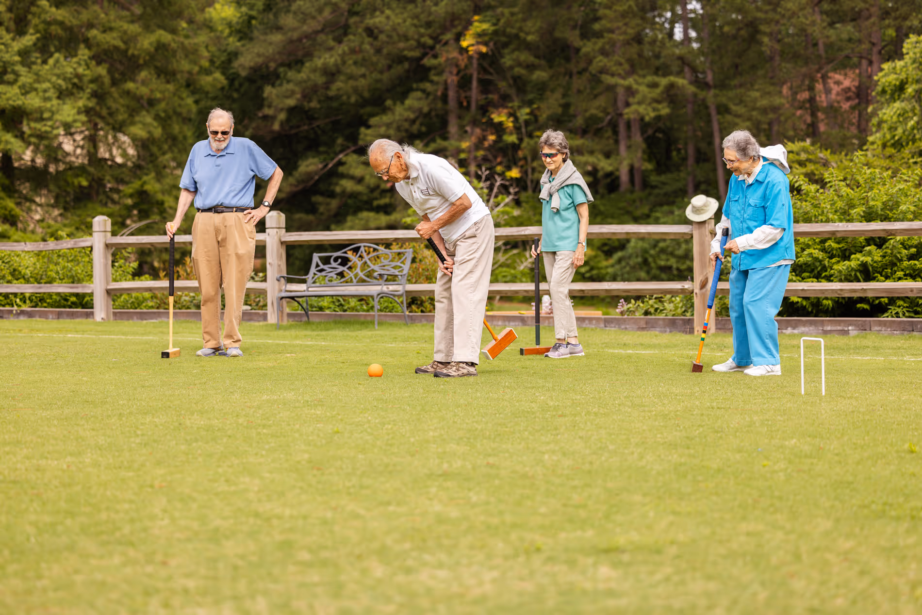 Four people playing croquet on grass, each aiming to hit balls with mallets toward hoops.