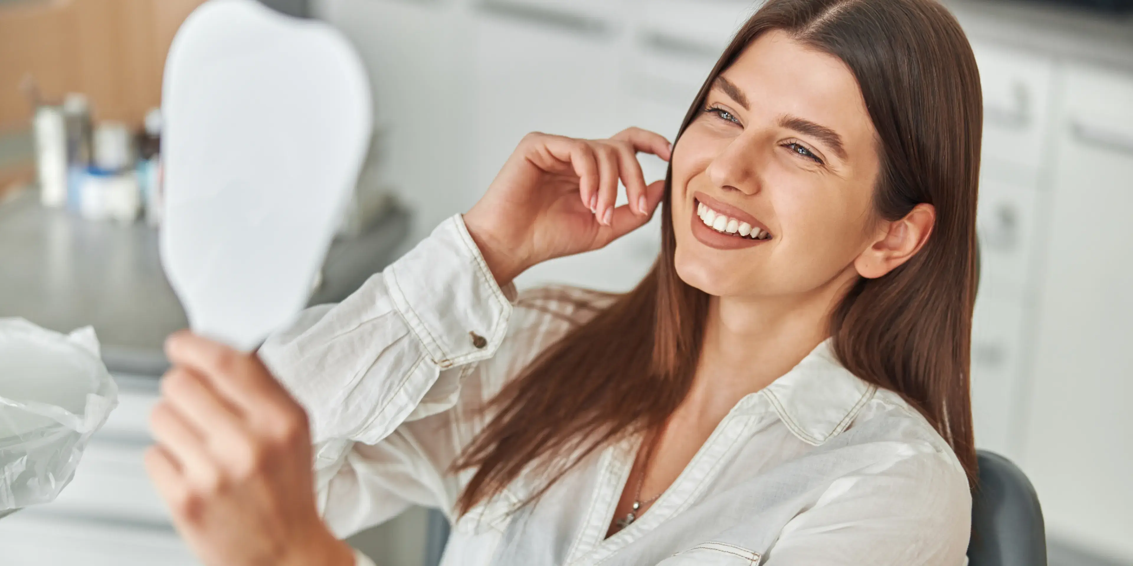 A woman smiles while looking at herself in a handheld mirror.
