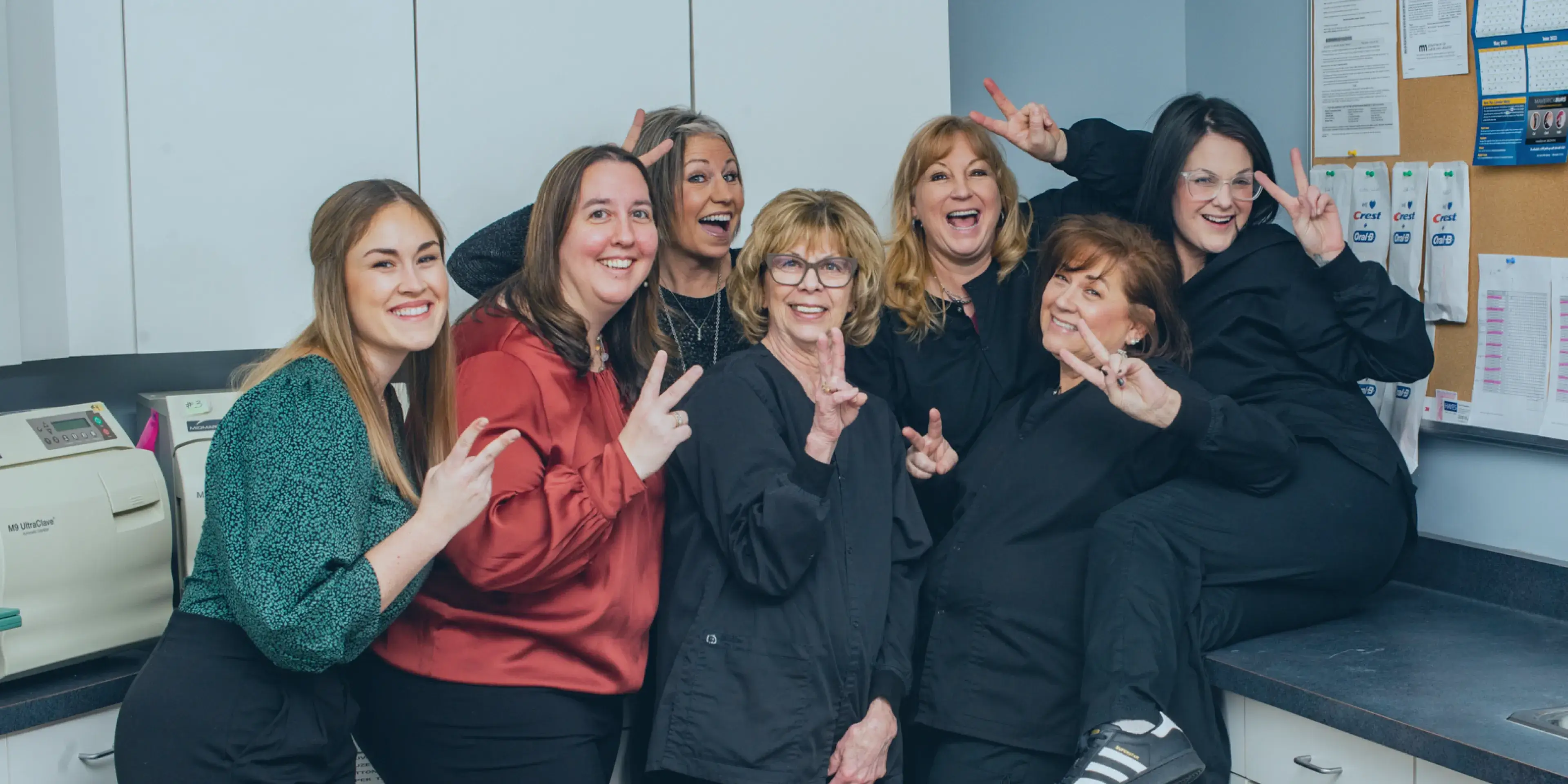Eight people smiling and posing together in a room, many making peace signs with their hands.