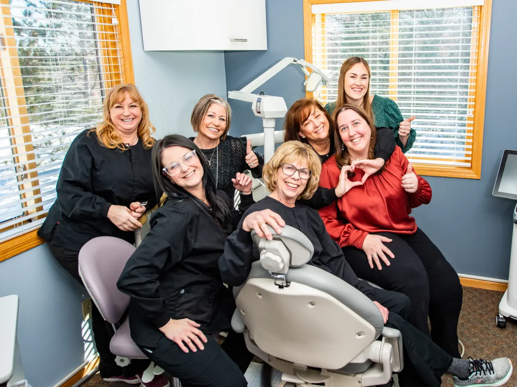 Seven people smiling and giving thumbs up in a dental office near a dental chair.