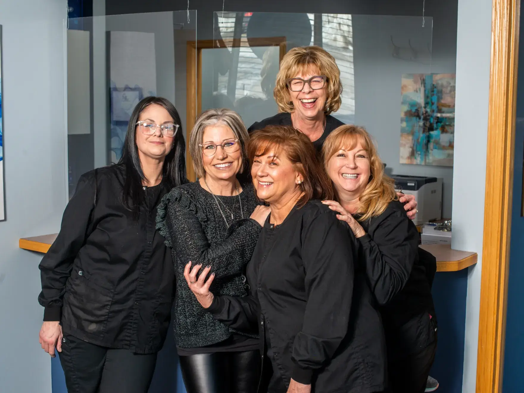 Five women stand together, smiling in an office setting.