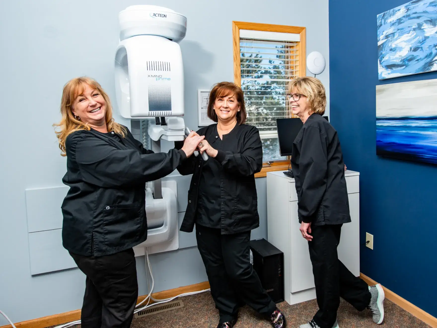 Three women in black scrubs smiling and holding hands in a medical office with equipment.