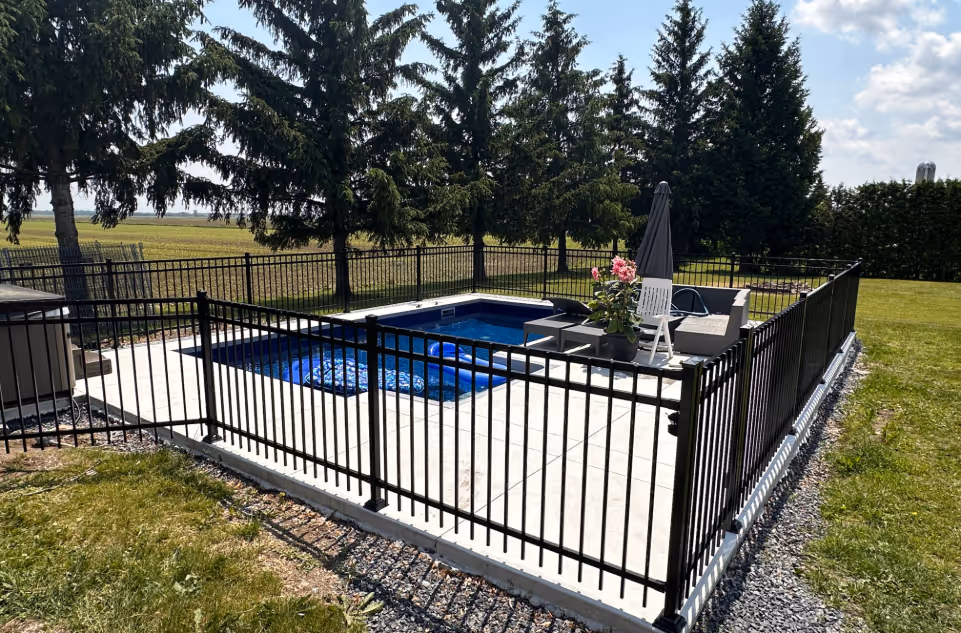 Piscine creusée avec un patio, entourée d'une clôture ornementale noire dans une cour avec des arbres en arrière-plan.