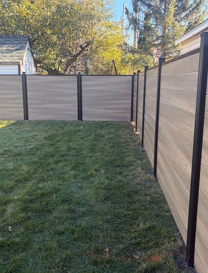 Clôture en panneaux taupe avec piliers noirs entourant une cour arrière herbeuse, arbres avec feuilles vertes en arrière-plan.